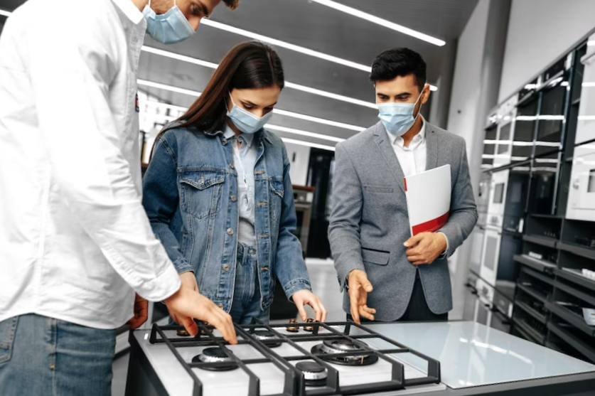 Shop assistant in hypermarket shows new model of gas stove to young couple all wearing medical masks