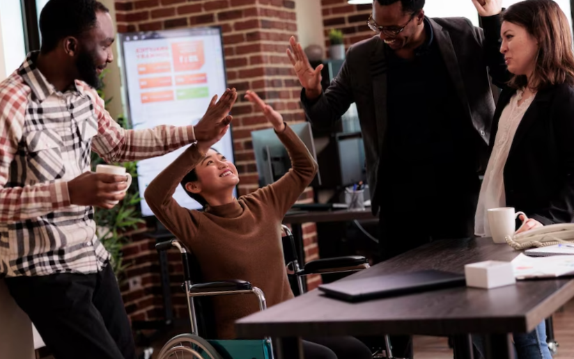 Multiethnic group of people having fun at work on break, laughing and enjoying conversation with paralyzed employee. woman living with physical chronic disability in wheelchair at job.
